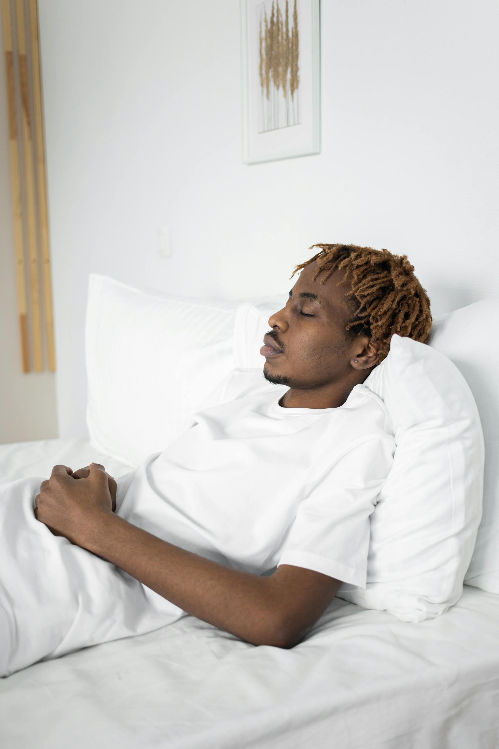 African American man sleeping peacefully on a white bed indoors.