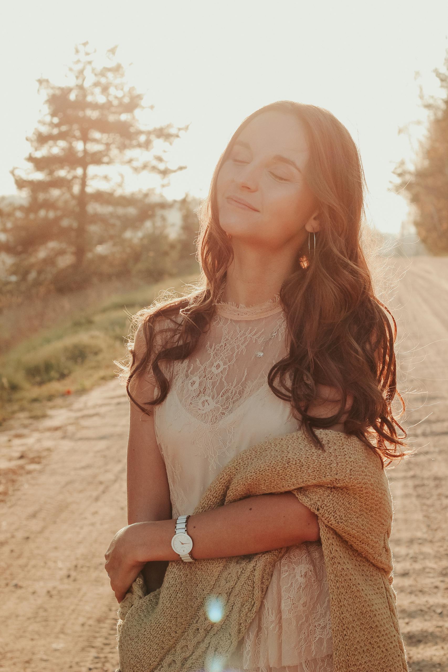 Young woman in lace dress and sweater basks in warm outdoor sunlight, embodying elegance and serenity.